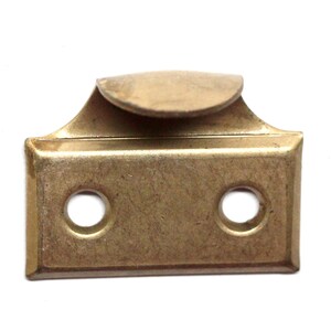 Puede incluir: Un pestillo de ventana de latón con dos agujeros y una parte superior curva. El pestillo tiene una forma rectangular con esquinas redondeadas.