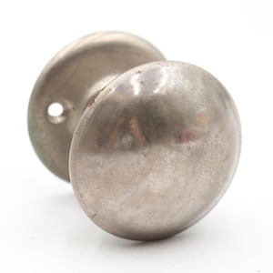 Op de afbeelding: Een ronde, zilverkleurige metalen deurknop met een glad, gepolijst oppervlak. De knop is voorzien van een ronde achterplaat met een zichtbaar schroefgat, wat een eenvoudig en functioneel ontwerp met een reflecterende, licht verweerde afwerking toont.