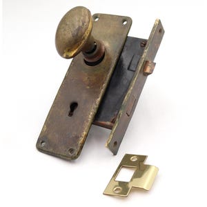 May include: Antique brass door hardware set, including a doorknob, faceplate with a keyhole, and a strike plate. The hardware shows signs of age and wear, with a tarnished finish. The set is designed for a vintage or rustic aesthetic.