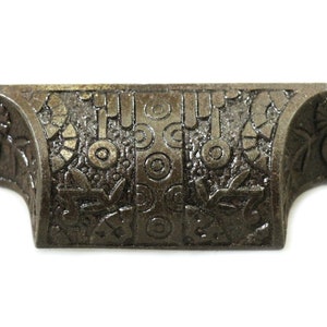 May include: A cast iron drawer pull with an ornate design featuring circles, lines, and floral motifs. The pull is a dark brown color and has a slightly curved shape.