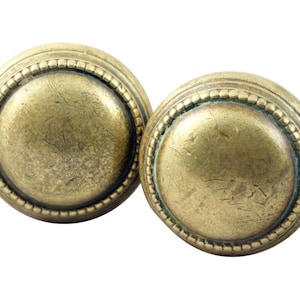 May include: Two antique brass cabinet knobs. Each knob has a circular design with a raised center and a beaded edge. The brass has a warm, aged patina, suggesting vintage or retro style. The knobs are set against a white background.