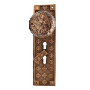 May include: Antique bronze door knob and backplate. The round knob and rectangular backplate feature an ornate floral pattern. The backplate has two keyholes and a small, rectangular keyhole cover.