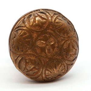 Może przedstawiać: Okrągły, złoty metalowy guzik z ozdobnym wzorem. Guzik ma wypukły, okrągły wzór z sześcioma sekcjami, z których każda zawiera inny motyw kwiatowy.