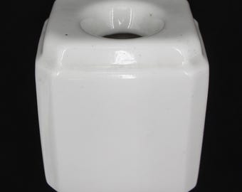 May include: A white, octagonal ceramic object with a circular opening at the top. The object has a glossy finish and is set against a black background. The design is simple and geometric.