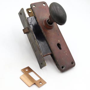 May include: Antique door hardware set featuring a reddish-brown door plate with a black oval doorknob. Includes a matching latch and a separate strike plate, all showing signs of age and wear. Ideal for restoration projects.