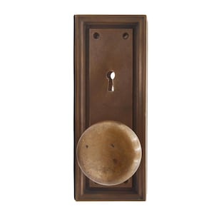 May include: A vintage bronze door handle set. The rectangular backplate features a keyhole and two screw holes. A round, aged doorknob is attached. The hardware has a weathered appearance, suggesting age and use.