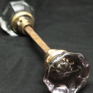 May include: Antique door knob with clear faceted glass knobs, brass accents, and a rusty metal rod. The door hardware is set against a dark background.
