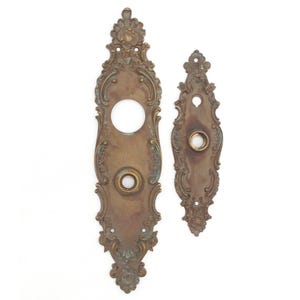 May include: Two antique brass door plates with ornate floral and scroll designs. The larger plate has a circular opening and a smaller button, while the smaller plate has a button. Both have a weathered patina.