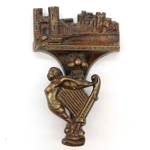 May include: A gold-toned metal souvenir featuring a harp and a depiction of Conwy Castle and Bridge. The inscription reads "Conwy Castle and Bridge".