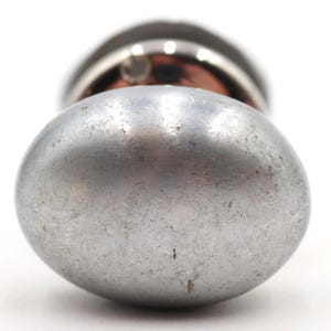 Op de afbeelding: Close-up van een zilverkleurig, ovaalvormig object met een getextureerd oppervlak. Het object heeft een metallic uitstraling en staat tegen een witte achtergrond. Het bovenste deel van het object is donker.