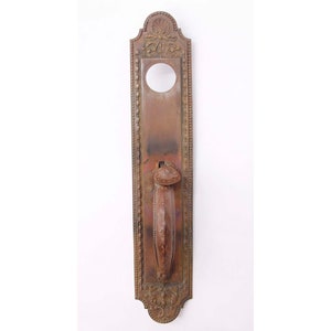 May include: Antique brass door handle with ornate details and a keyhole. The handle is attached to a rectangular plate with a decorative border.