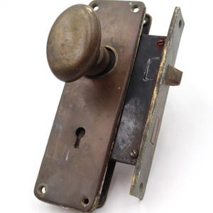 May include: Antique bronze door hardware set, featuring an oval doorknob and a rectangular backplate with a keyhole. The set includes a matching strike plate. The metal has a weathered appearance.