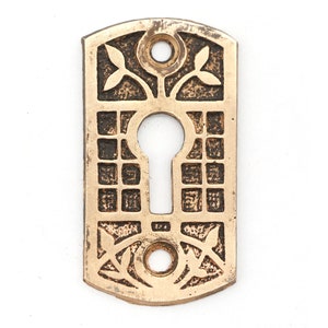 Może przedstawiać: Okładzina otworu na klucz wykonana z mosiądzu o skomplikowanym wzorze z geometrycznym wzorem i motywami liści. Okładzina ma kształt prostokąta ze zaokrąglonymi narożnikami.