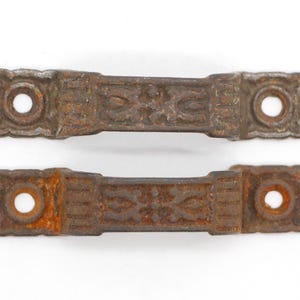 Może przedstawiać: Dwa antyczne, zardzewiałe metalowe uchwyty do szuflad z ozdobnymi wzorami. Uchwyty są prostokątne ze zaokrąglonymi końcami i mają dwa otwory na śruby.