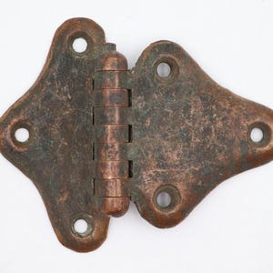 May include: A vintage copper-colored hinge with a decorative shape. The hinge has a central barrel with multiple segments and two flared, diamond-shaped leaves, each with three screw holes. The metal has a mottled, aged appearance.
