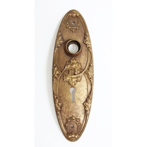 May include: An oval-shaped brass keyhole cover with an intricate floral design. The cover has a keyhole in the center and a small round hole at the top.