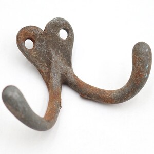 May include: A double-pronged metal wall hook featuring a weathered, rusted brown and gray finish. The mounting plate at the top includes two circular screw holes, and the two curved hooks extend outward in opposite directions from the center.