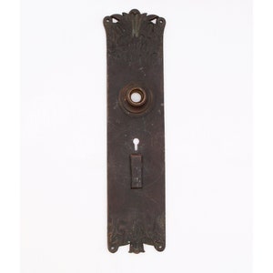 May include: A rectangular, antique bronze door plate with ornate floral and leaf designs. The plate features a keyhole, a door knob opening, and a rectangular latch. The decorative plate is a vintage architectural element.