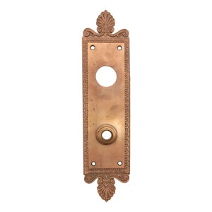 May include: A brass door plate with an ornate design and a keyhole. The plate is rectangular and has a decorative border.