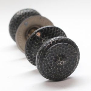 May include: Three black metal door knobs with a hammered finish. The knobs are round and have a raised edge. The knobs are arranged in a stack, with the largest knob on the bottom.
