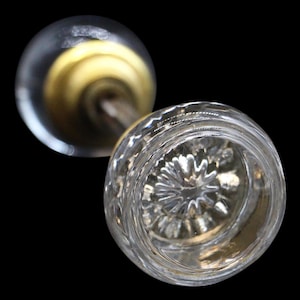 Può includere: Due pomelli per porte in vetro trasparente con un design decorativo e strutturato. Un pomello è a fuoco, mostrando un motivo dettagliato, mentre l'altro è leggermente sfocato. L'hardware ha uno stelo color ottone ed è posto su uno sfondo nero.