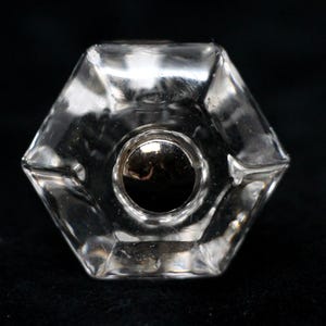 Puede incluir: Un tapón de botella de vidrio transparente con forma hexagonal y una parte superior de metal plateado.