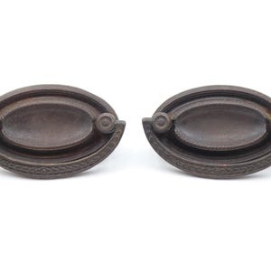Puede incluir: Dos tiradores de cajón antiguos de color bronce. Cada tirador tiene forma ovalada con un área central empotrada y detalles decorativos en los bordes. Pequeños acentos circulares en cada extremo.