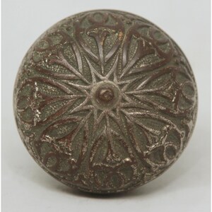 May include: A round, antique-style metal knob with an intricate floral design. The knob is primarily silver with reddish-brown accents, and the detailed pattern radiates from a central point. This decorative hardware is suitable for furniture or cabinetry.