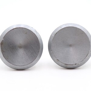 Puede incluir: Dos objetos circulares de metal plateado con acabado cepillado. Los objetos son redondos con una parte superior plana y un borde ligeramente redondeado. Los objetos están sobre un fondo blanco.