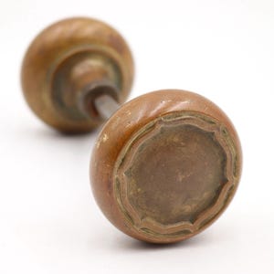 May include: Close-up of two antique brass door knobs. The knobs have a rounded shape with a decorative edge. The metal has a warm, aged patina, suggesting vintage craftsmanship. The knobs are connected by a metal rod.