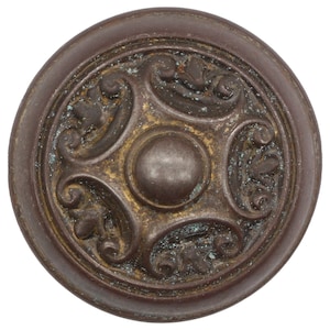 May include: A round, ornate, bronze-coloured door knob with a raised, circular centre and a decorative, floral design.