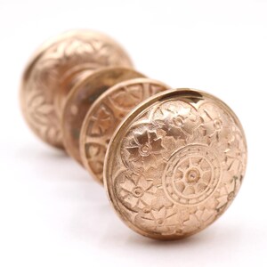 May include: A set of three round, metallic, rose-gold colored drawer knobs arranged in a row. Each knob features a detailed, embossed floral pattern across its circular face, with a central circular motif surrounded by stylized flower petals.