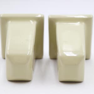 May include: Two cream-coloured ceramic towel hooks. Each hook features a rectangular base and a curved, rectangular arm. The hooks are placed side-by-side against a white backdrop. The ceramic has a glossy finish.