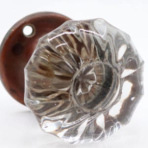 May include: A clear, faceted glass doorknob with a bronze-colored backplate. The doorknob has a detailed, octagonal design, reflecting light. The backplate is round with a single hole. This vintage hardware adds a classic touch to any door.