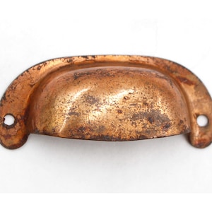 May include: A vintage copper drawer pull with a rounded, slightly curved shape. The pull has a worn, aged patina and two small holes for mounting.