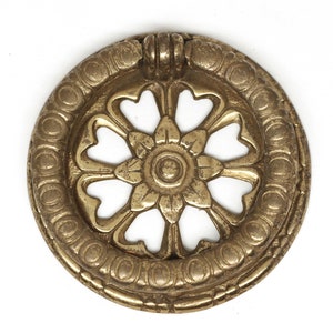 Puede incluir: Un tirador de armario circular de color latón con un diseño floral. El centro presenta una forma de flor, rodeada de recortes en forma de corazón y un anillo exterior decorativo. El tirador tiene un pequeño asa en la parte superior.