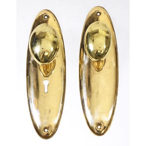 May include: Two oval-shaped brass door knobs with a keyhole and a round knob. The knobs are polished and have a shiny finish.