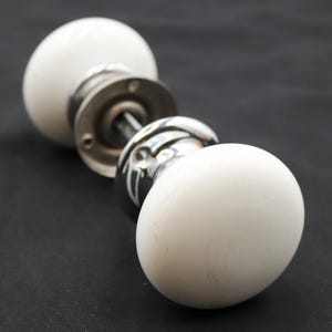 May include: A door knob set featuring two smooth, round, white ceramic handles attached to a central metal spindle. The handles are connected to polished chrome circular base plates, each containing two small screw holes for mounting purposes.