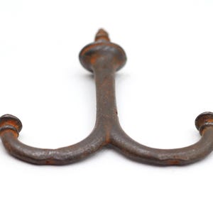 May include: A double-pronged metal wall hook with a dark, weathered, and rusted finish. The hook features a central vertical stem topped with a rounded finial and two curved arms extending outward, each ending in a small, rounded decorative knob.