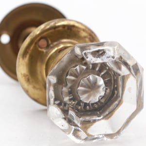 May include: Antique door knob with a clear, faceted glass handle and a tarnished brass base. The knob has an octagonal shape and a detailed, starburst design on the inside. The brass base shows signs of age and wear.