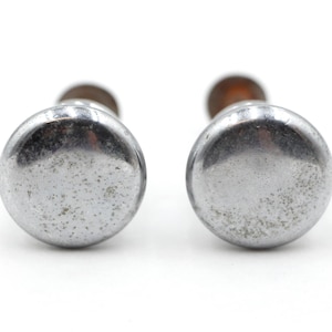 May include: Two vintage, silver-toned cabinet knobs with a round, slightly domed design. The knobs show signs of wear, with a mottled appearance. The stems are a darker color, and the background is white.