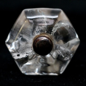 Puede incluir: Pomo de armario de cristal transparente y facetado con un centro oscuro y redondo. El pomo tiene forma hexagonal y está sobre un fondo negro. Este herraje de estilo vintage es adecuado para la restauración de muebles o para añadir un toque único a armarios y cajones.