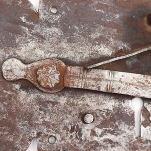 May include: Close-up of a weathered, antique metal lock mechanism. The lock features a keyhole, a lever, and a decorative handle with a starburst design. The metal has a rusty brown and silver patina, suggesting age and use. The lock is likely from a door or chest.