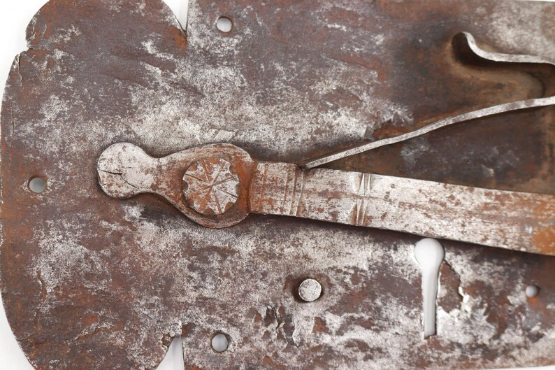 May include: Close-up of a weathered, antique metal lock mechanism. The lock features a keyhole, a lever, and a decorative handle with a starburst design. The metal has a rusty brown and silver patina, suggesting age and use. The lock is likely from a door or chest.