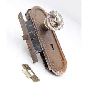 May include: An antique door knob set with a clear, faceted glass knob and a tarnished, bronze-colored backplate. The set includes a black metal lock mechanism and a separate strike plate.