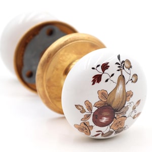 May include: White ceramic door knobs with a gold-colored base and a fruit design. The fruit design includes a pear, apples, and leaves in shades of brown, red, and tan. The knobs are for a door or cabinet.