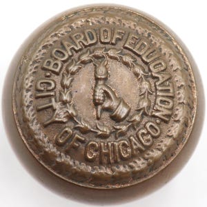 May include: A bronze-colored, round object with the text "CITY BOARD OF EDUCATION OF CHICAGO" encircling a central emblem of a hand holding a torch. The object has a slightly domed shape and a detailed, textured surface.