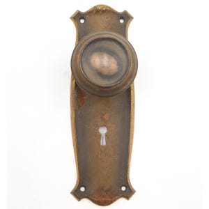 May include: Antique bronze door knob and backplate. The door knob is round and the backplate is rectangular with decorative edges. The finish has a weathered appearance, showing signs of age and use. The keyhole is centered on the backplate.