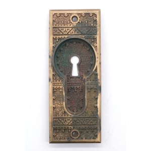 Może przedstawiać: Okładzina otworu na klucz wykonana z mosiądzu z ozdobnym wzorem. Okładzina jest prostokątna i ma otwór na klucz w środku. Wzór jest skomplikowany i zawiera różne wzory.