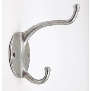 May include: A vintage, silver-colored metal coat hook with a double hook design. The hook is mounted on a round base with two screw holes for easy installation. The metal has a slightly aged appearance, suggesting it is an antique or vintage item.